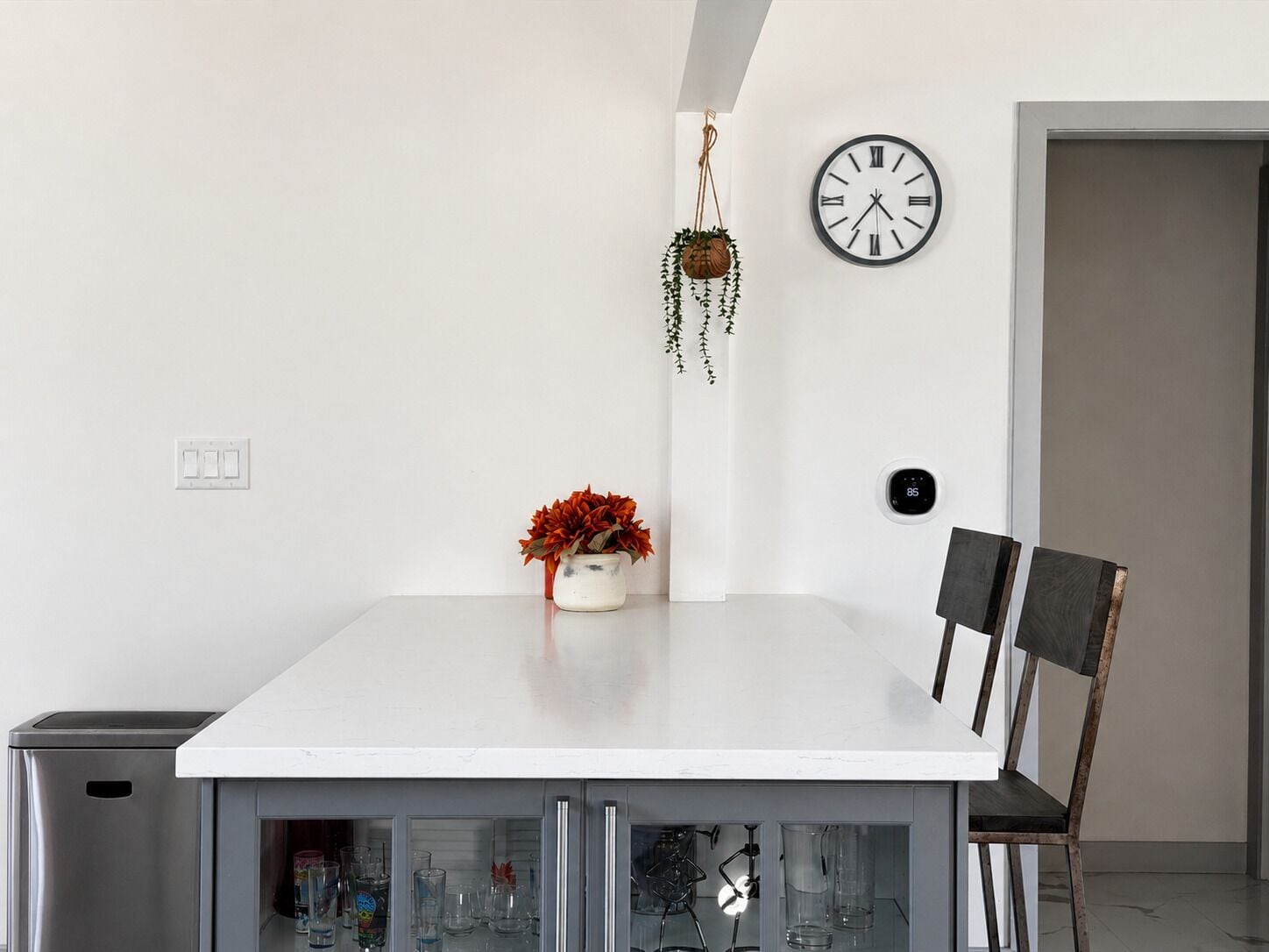 A modern dining area with a white table, two chairs, a hanging plant, a wall clock, and a thermostat.