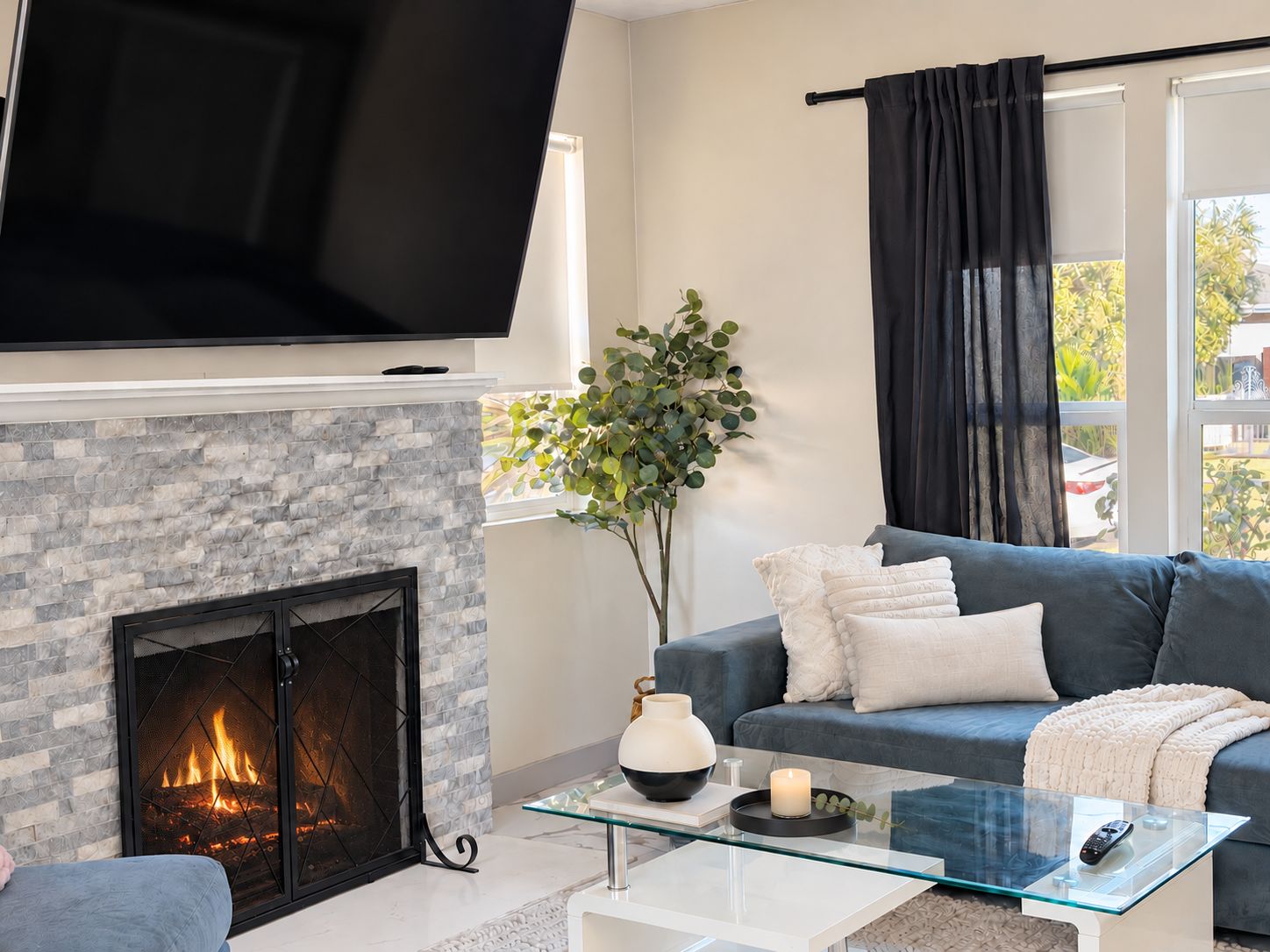 A living room with a fireplace, a flat-screen TV mounted above it, a sofa, a glass coffee table, and a window with a view of greenery outside.