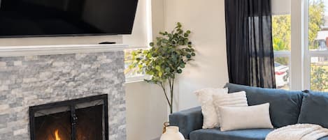 A living room with a fireplace, a flat-screen TV mounted above it, a sofa, a glass coffee table, and a window with a view of greenery outside.