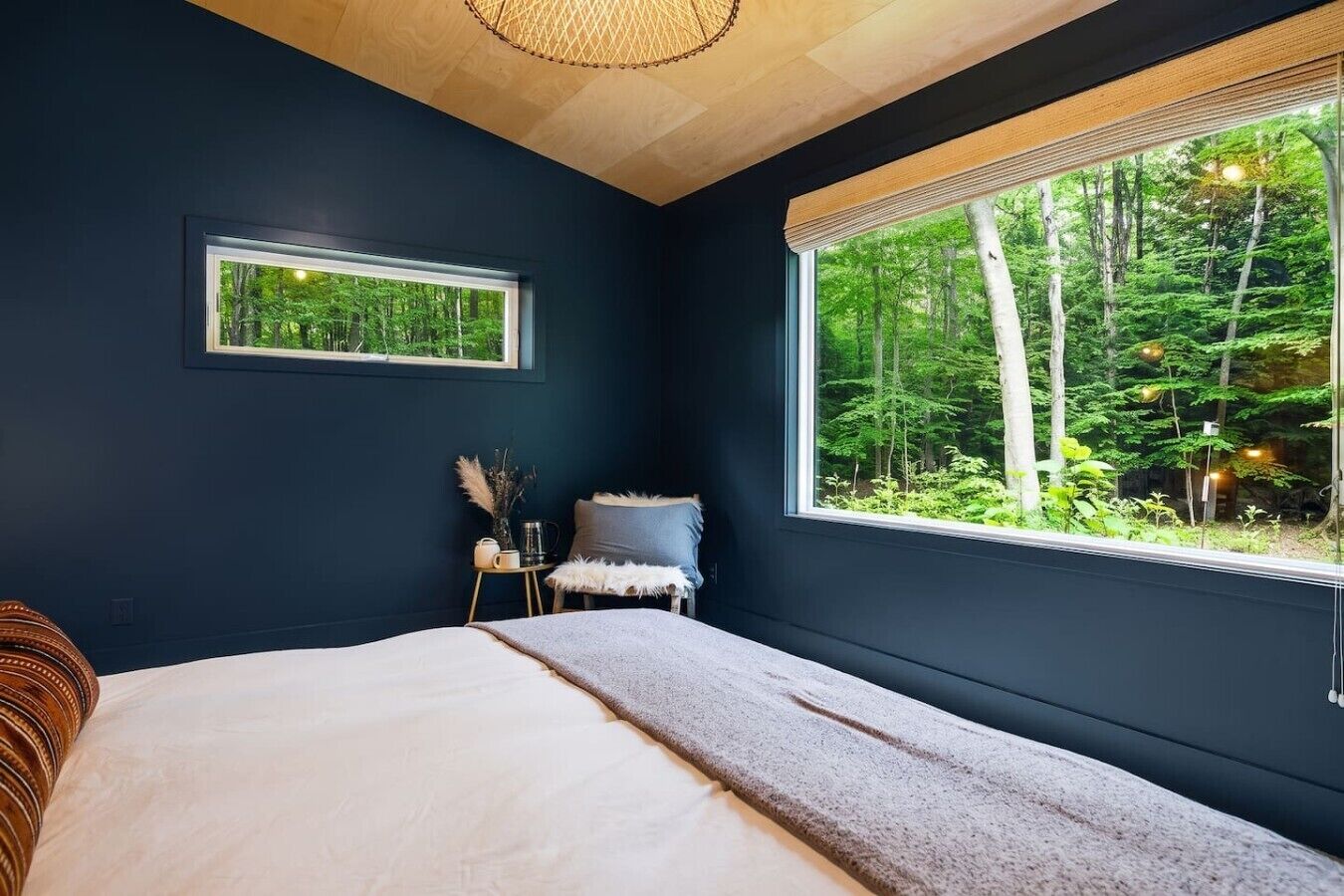 A bedroom with a large window, a bed, a bedside table with a lamp, and a view of a forest.