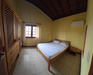 A room with a bed, a wooden wardrobe, a window with curtains, and an air conditioning unit. - Charming 2-Bedroom Apartment in Enchanting Porto Seguro with AC Comfort (Porto Seguro)