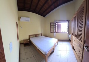A bedroom with a bed, a nightstand, a wardrobe, and a window with shutters. - Charming 2-Bedroom Apartment in Enchanting Porto Seguro with AC Comfort (Porto Seguro)