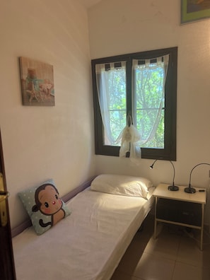Ein Einzelbett mit weißer Matratze und einem Kopfkissen mit einer Zeichentrickfigur, ein Nachttisch mit Lampe sowie ein Fenster mit durchscheinenden Vorhängen.
