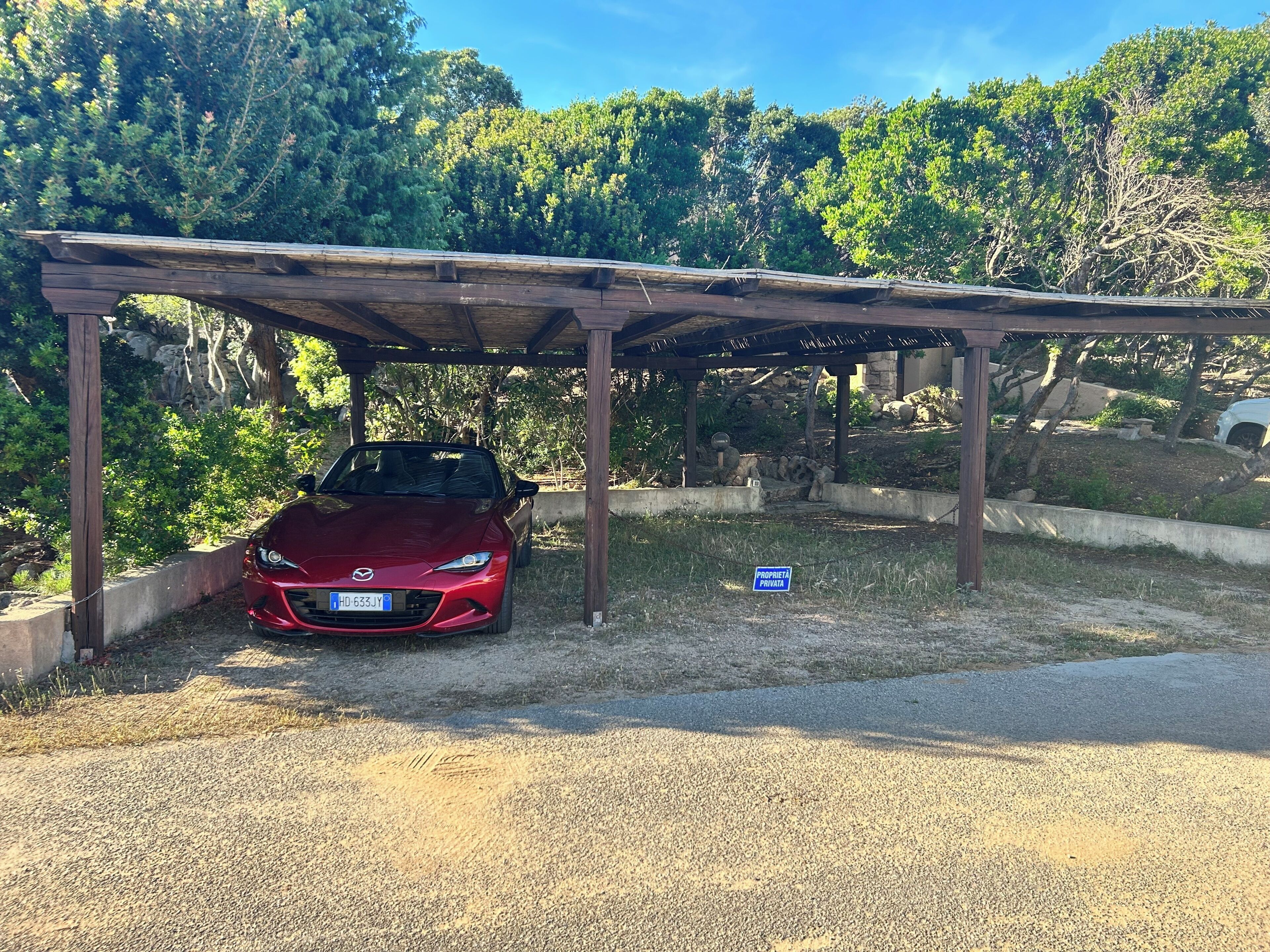Un coche rojo estacionado bajo una pérgola de madera, con un entorno natural de árboles y cielo despejado.