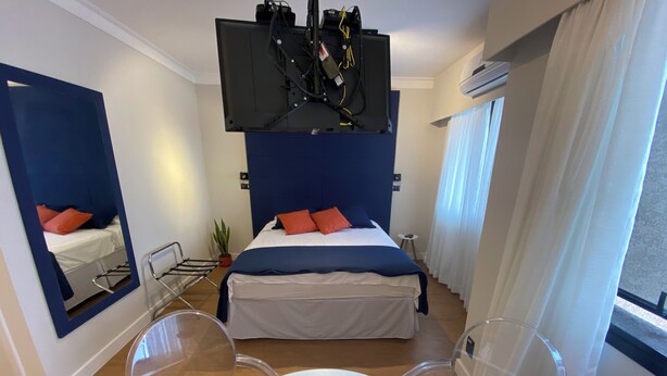 Ein modernes Hotelzimmer mit Bett, Spiegel, Tisch mit Blumenvase und einem am Decken befestigten Fernseher.