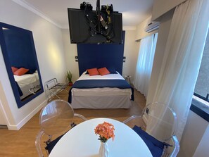 Ein modernes Hotelzimmer mit Bett, Spiegel, Tisch mit Blumenvase und einem am Decken befestigten Fernseher.