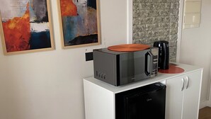Eine kompakte Küchenzeile mit Mikrowelle, Kaffeemaschine und kleinem Kühlschrank. An der Wand befinden sich zwei abstrakte Gemälde, ein transparenter Stuhl und ein Tisch mit Blumen.'