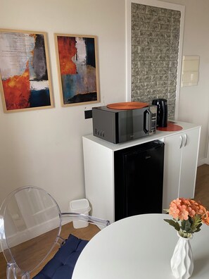 A compact kitchenette with a microwave, coffee maker, and a small refrigerator. There are two abstract paintings on the wall, a clear chair, and a table with a vase of flowers. - Functional apartment opposite Congonhas Airport. (São Paulo)