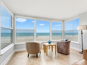 A room with large windows overlooking a beach, two chairs, a small table, and a lamp. - Madeira Vista 202 (Madeira Beach)