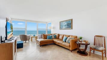 A living room with a large window overlooking the sea, a brown leather sofa, a wooden side table, and a framed painting on the wall.