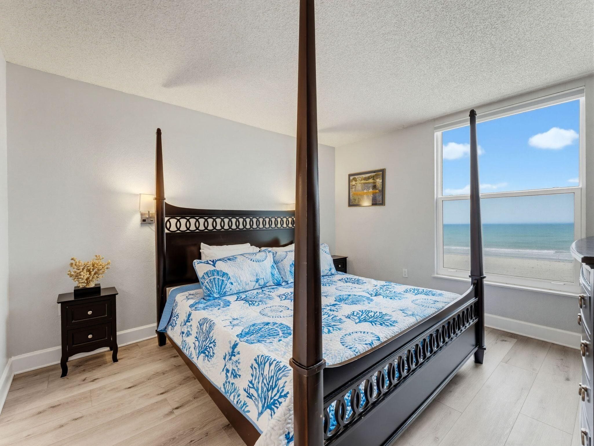 A four-poster bed with a blue and white floral bedspread, a nightstand with a vase of dried flowers, a painting on the wall, and a view of the sea through a large window.