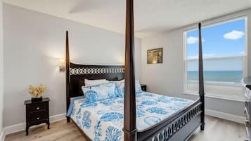 A four-poster bed with a blue and white floral bedspread, a nightstand with a vase of dried flowers, a painting on the wall, and a view of the sea through a large window.