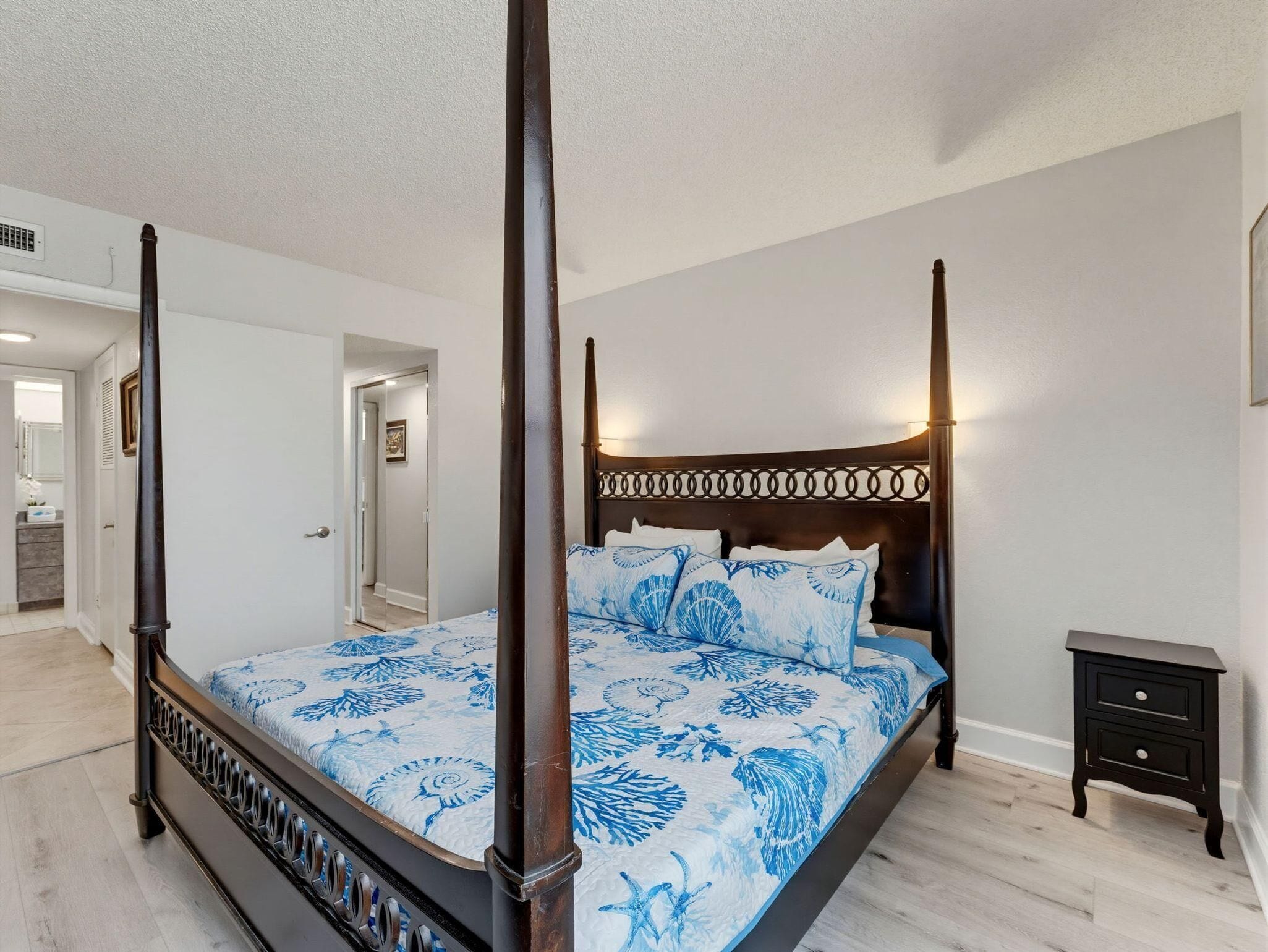 A four-poster bed with a floral bedspread, a nightstand, and a hallway leading to another room.