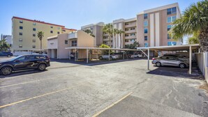 Un estacionamiento con varios coches, una estructura cubierta para vehículos y un edificio con varios pisos y balcones.