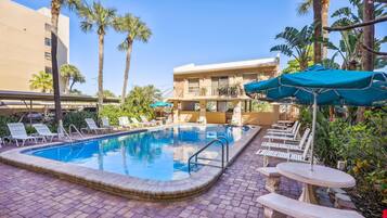 A swimming pool with lounge chairs and umbrellas, surrounded by palm trees and a building with balconies.