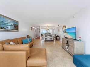 A modern living room with a leather sofa, a dining area with a chandelier, and a TV displaying a beach scene. - Madeira Vista 202 (Madeira Beach)