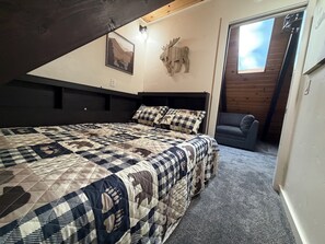 Une chambre à coucher avec un grand lit, un plafond en bois, une tête de wapiti accrochée au mur, un tableau encadré et une fenêtre.