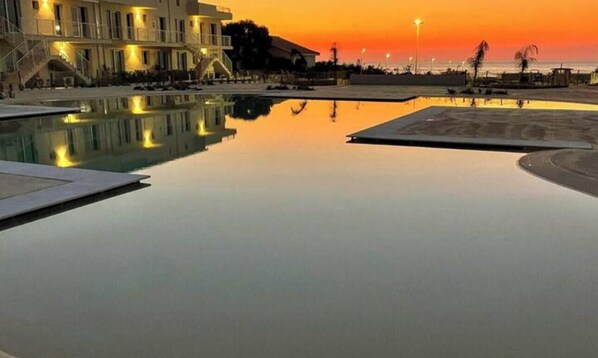 A swimming pool with a reflection of a building and a sunset in the background. - Largest 2 Bedroom Oceanview Rooftop Apartment in Borto 84 (Sicilia)