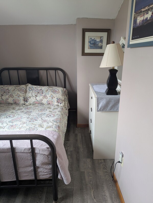 A bedroom with a bed, a bedside table with a lamp, a framed picture on the wall, and a dresser.