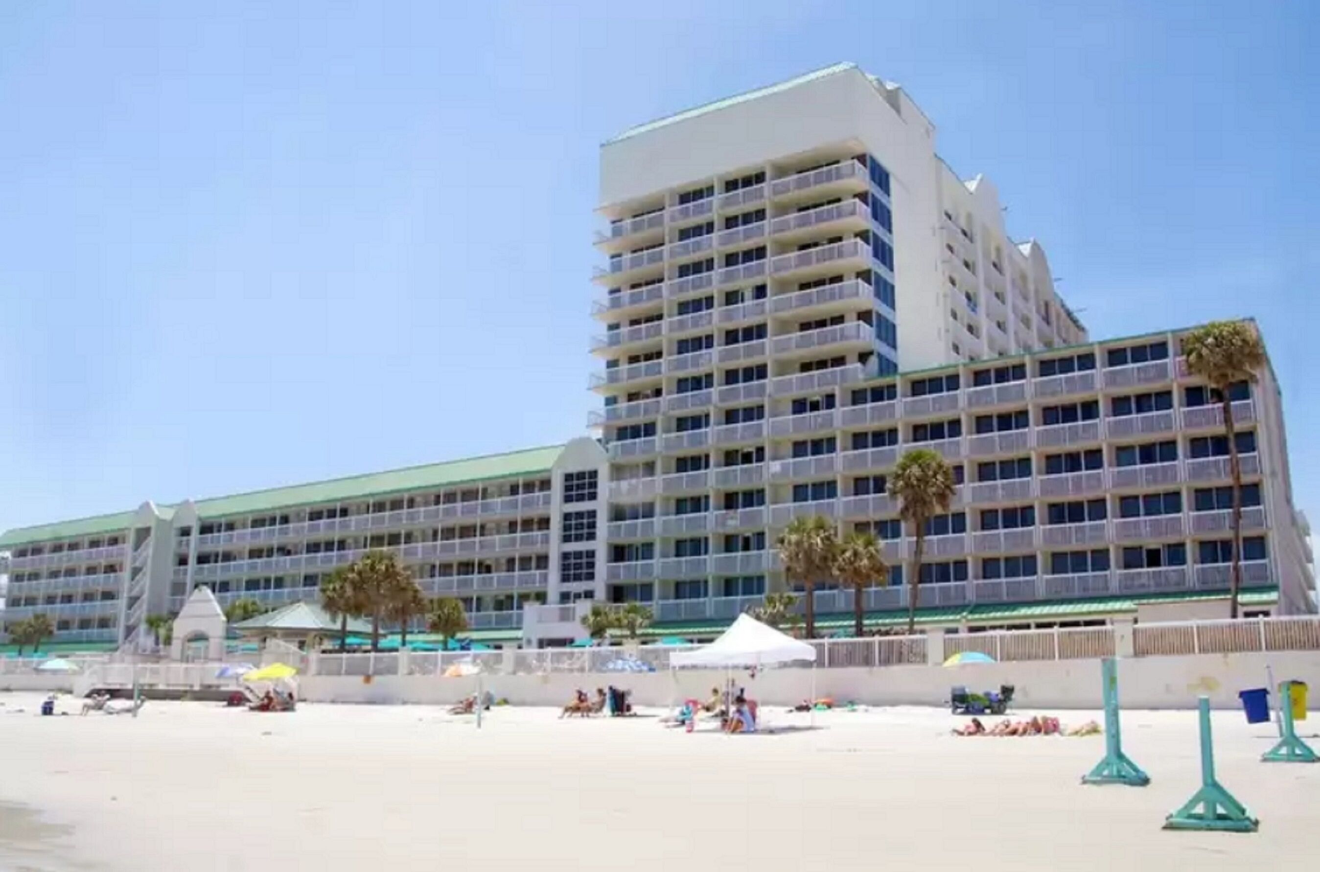 Et stort hotel med flere etager og altaner, en strand i forgrunden og palmer.