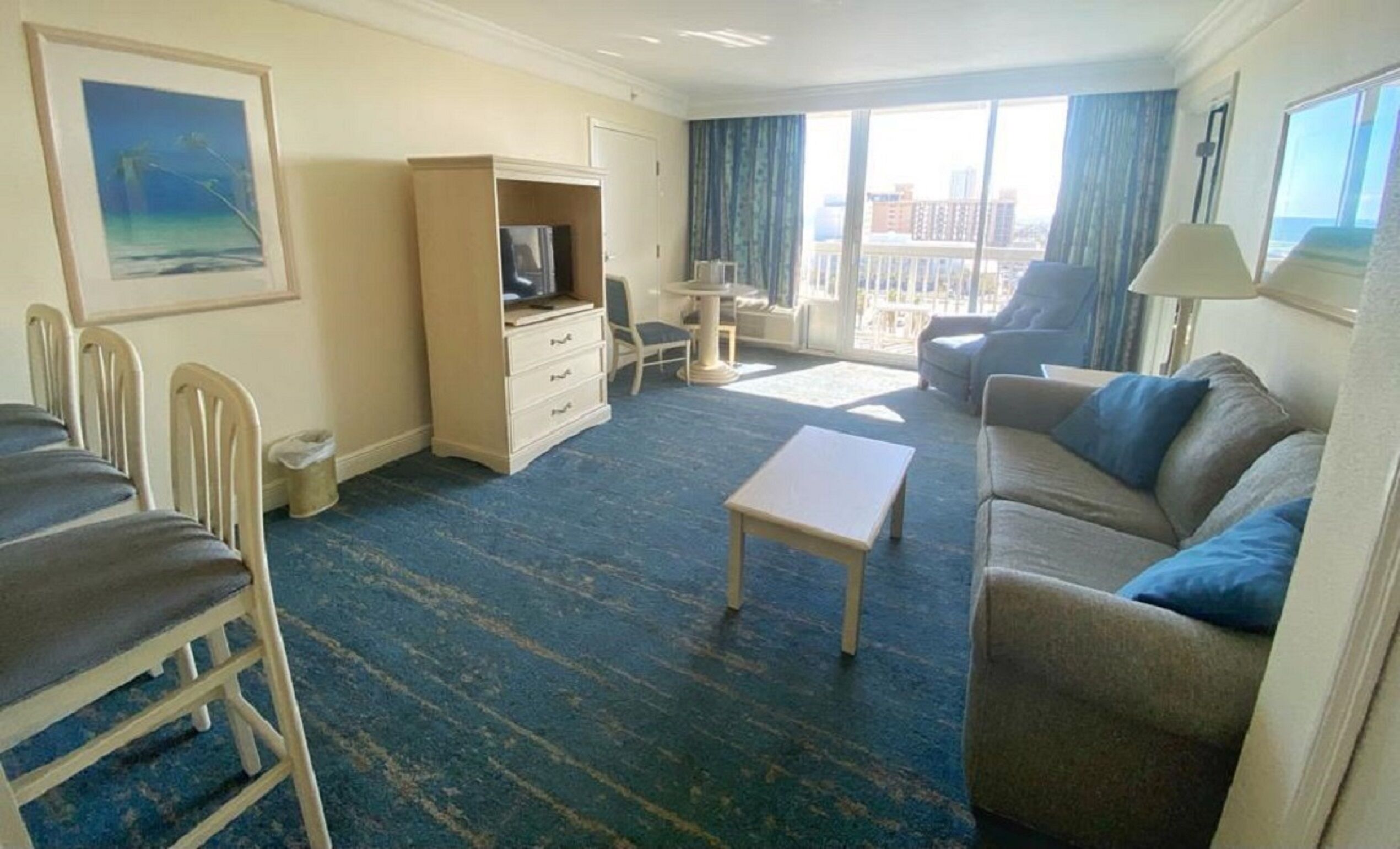 Quarto de hotel com sofá, mesa pequena, televisão, cadeira, escrivaninha e varanda com cortinas.
