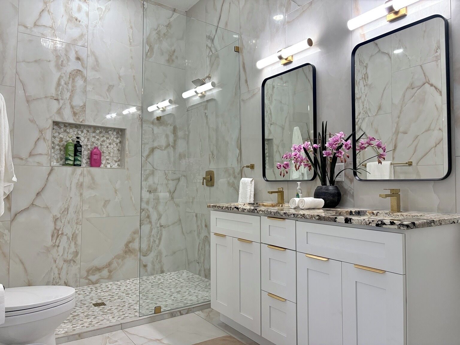 Un baño moderno con paredes de mármol, una cabina de ducha de vidrio, un tocador con tiradores dorados y un espejo con marco negro.