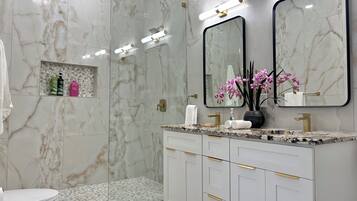 Un baño moderno con paredes de mármol, una cabina de ducha de vidrio, un tocador con tiradores dorados y un espejo con marco negro.