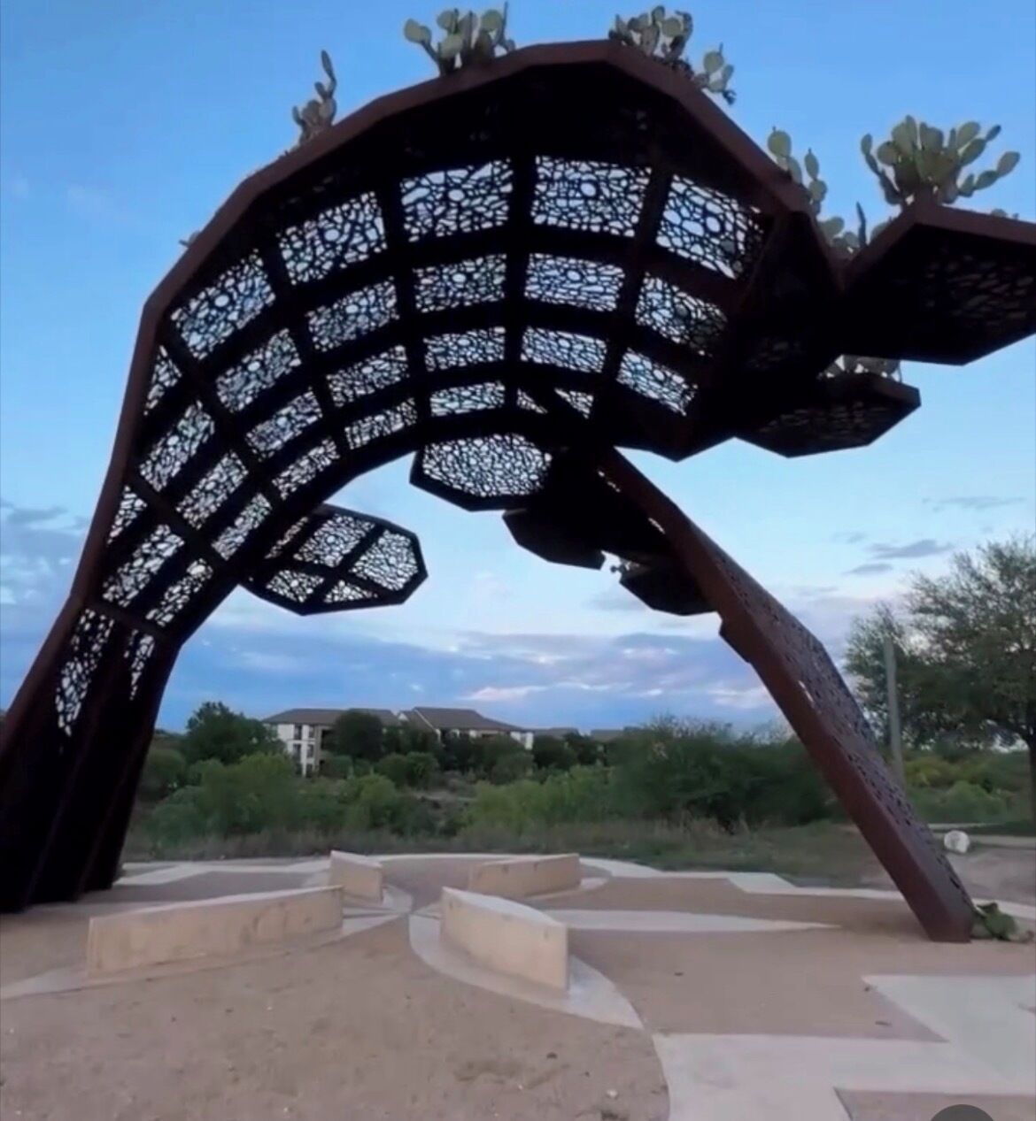 Una escultura de metal con un diseño de celosía, ubicada sobre una plataforma de concreto con escalones que la rodean.