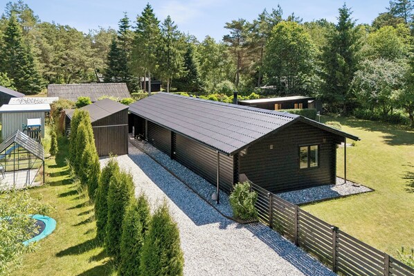 A wooden cabin with a gravel driveway, surrounded by trees and a fenced yard.
