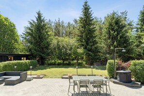 Een terras met een tafel en stoelen, omgeven door een verzorgde tuin met verschillende bomen en struiken.