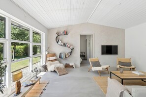 A spacious living room with large windows, a curved bookshelf, two wicker chairs, a flat-screen TV, and a coffee table.