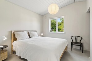 A bedroom with a bed, a chair, a bedside table, a lamp, and a window with a view of greenery.