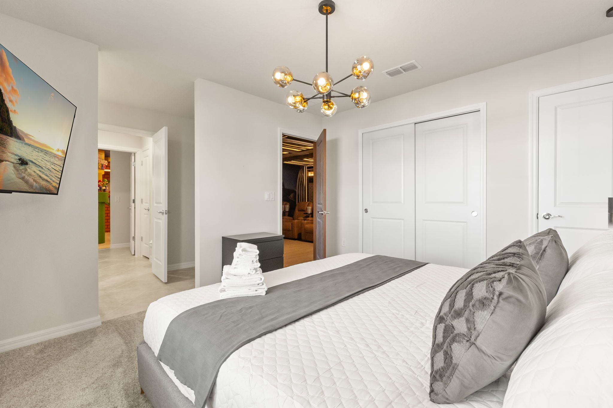 A bedroom with a bed, a bedside table, a wall-mounted TV, a ceiling light fixture, and a door leading to another room.