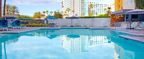 Una piscina con sillones de descanso y sombrillas, rodeada de palmeras y un hotel Marriott al fondo.
