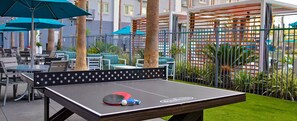 Una mesa de ping pong al aire libre con red, rodeada de sillas y mesas, con sombrillas y una cerca al fondo.