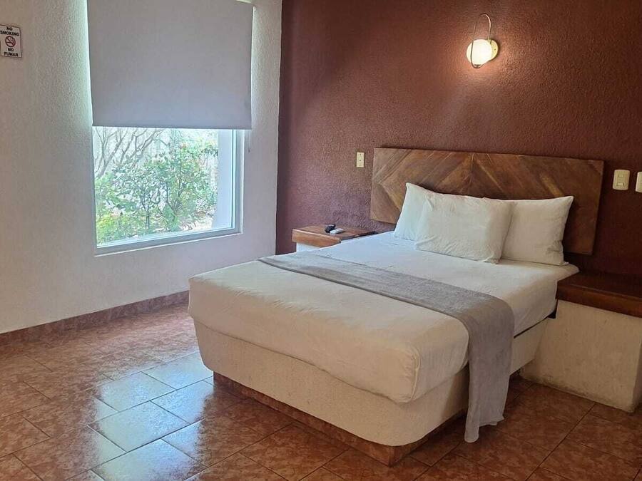 A bedroom with a bed, a nightstand, a window with a view, and a wall-mounted light fixture.