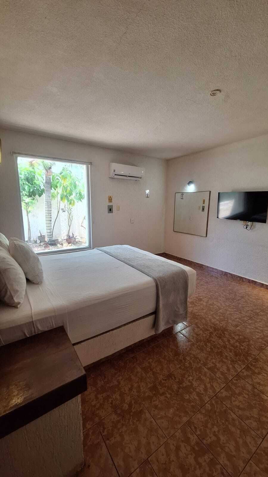 A bedroom with a bed, a window, an air conditioner, a flat-screen TV, and a tiled floor.