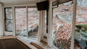 A room with large windows, a flat-screen TV, and a view of a rocky outdoor area with fallen leaves. - Glass Treehouse: waterfalls, boulders, hot tub, couples retreat (Banner Elk)