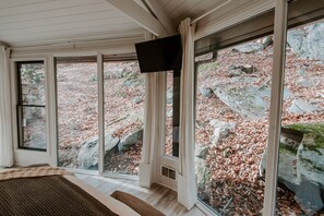 Ein Zimmer mit großen Fenstern, einem Flachbildfernseher und Blick auf eine felsige Landschaft mit herabgefallenen Blättern.
