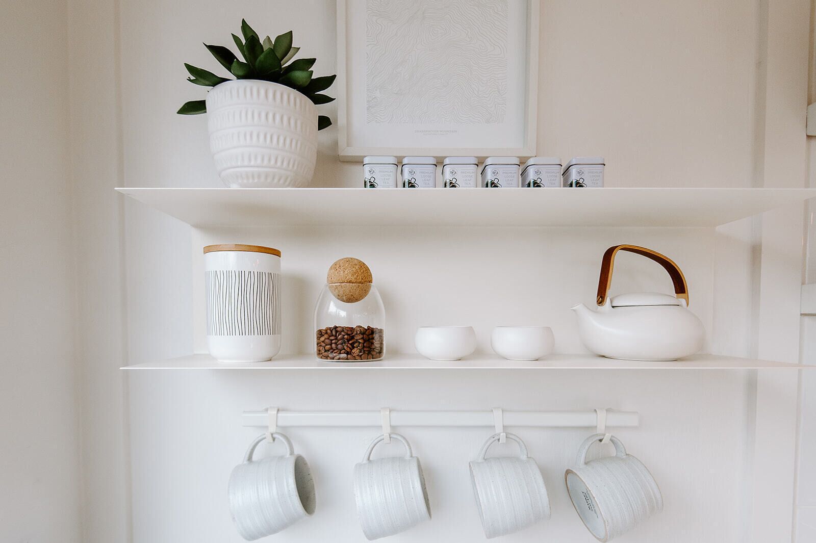 Een keukenschap met een plant in pot, een koffiemolen, een theepot en hangende mokken.