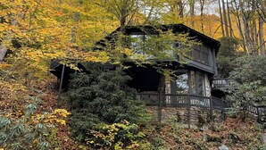 A cabin surrounded by autumn foliage, with a stone foundation and a wooden deck. - Glass Treehouse: waterfalls, boulders, hot tub, couples retreat (Banner Elk)