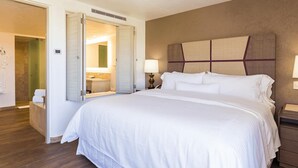 Hotellihuone, jossa on suuri sänky, yöpöytä, peili ja kylpyhuone, joka näkyy avoimesta ovesta.