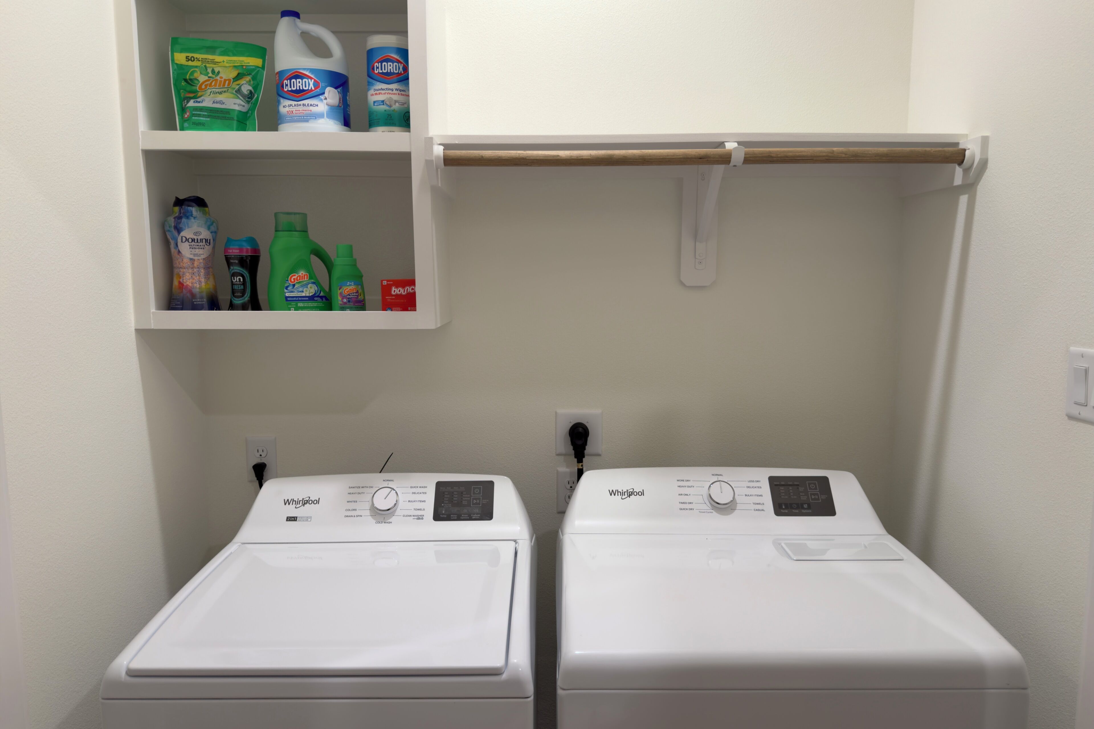 Two front-loading Whirlpool washers with a shelf above containing various cleaning products.