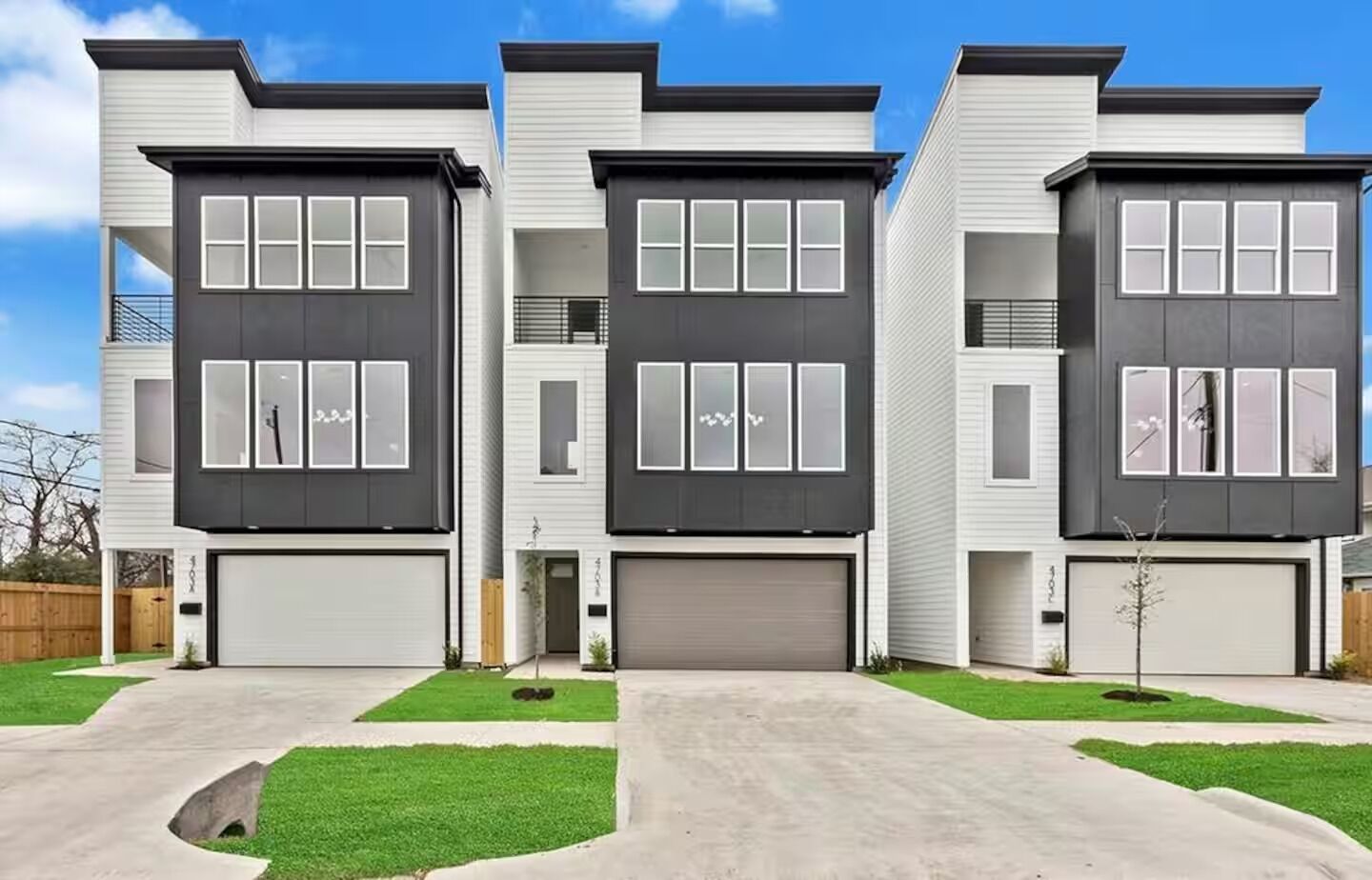 A modern three-story residential building with a garage on the ground floor, a small front yard, and a clear blue sky.