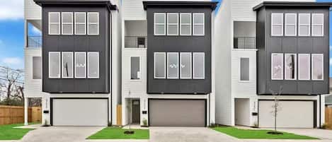A modern three-story residential building with a garage on the ground floor, a small front yard, and a clear blue sky.