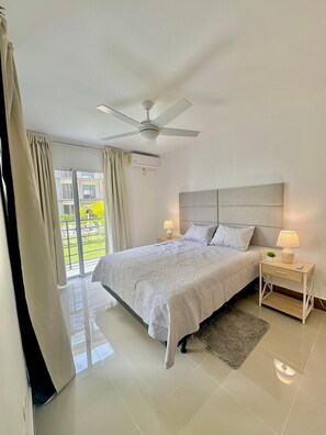 A bedroom with a bed, nightstands, a ceiling fan, and a view of greenery outside. - Golf, Beach, & Relax Punta Cana (Punta Cana)