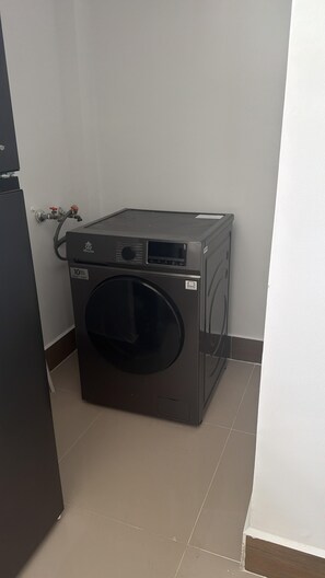 A compact, dark-colored washing machine in a corner of a room with a tiled floor and a white wall. - Golf, Beach, & Relax Punta Cana (Punta Cana)
