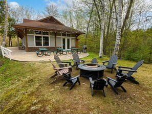 An outdoor seating area with a fire pit, surrounded by Adirondack chairs, and a covered pavilion with picnic tables in a wooded setting.