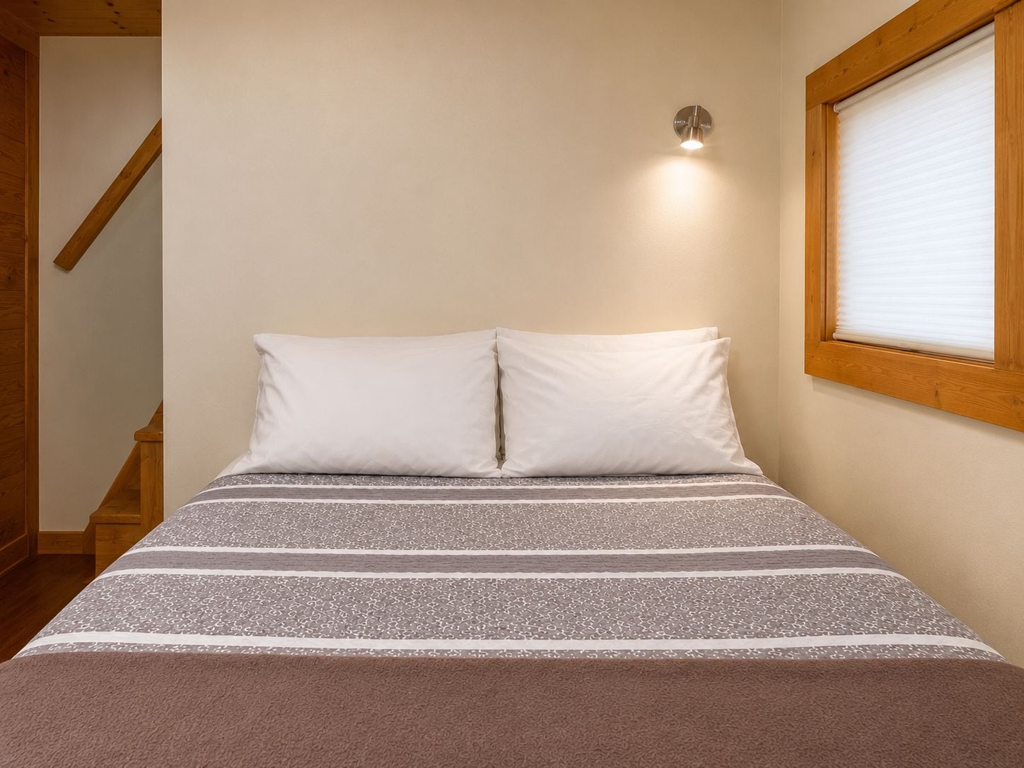 A bedroom with a bed, pillows, a window with blinds, a wall-mounted light, and a wooden staircase.
