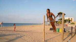 A beach with a pull-up bar and exercise equipment, a person doing a pull-up, and another person standing in the sand. - Cambrils-Salou Beach A (Cambrils)